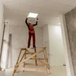 un operaio su un trabattello in legno mette un pannello di cartongesso sul controsoffitto sulla struttura
