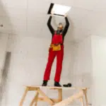 un operaio su un trabattello in legno mette un pannello di cartongesso sul controsoffitto sulla struttura