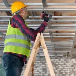operaio con caschetto giallo e gilet catarifrangente su una scala realizza una struttura per controsoffitto