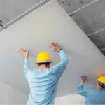 due operai con caschetto giallo mettono un pannello di cartongesso sulla struttura di un controsoffitto