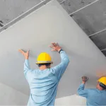 due operai con caschetto giallo mettono un pannello di cartongesso sulla struttura di un controsoffitto